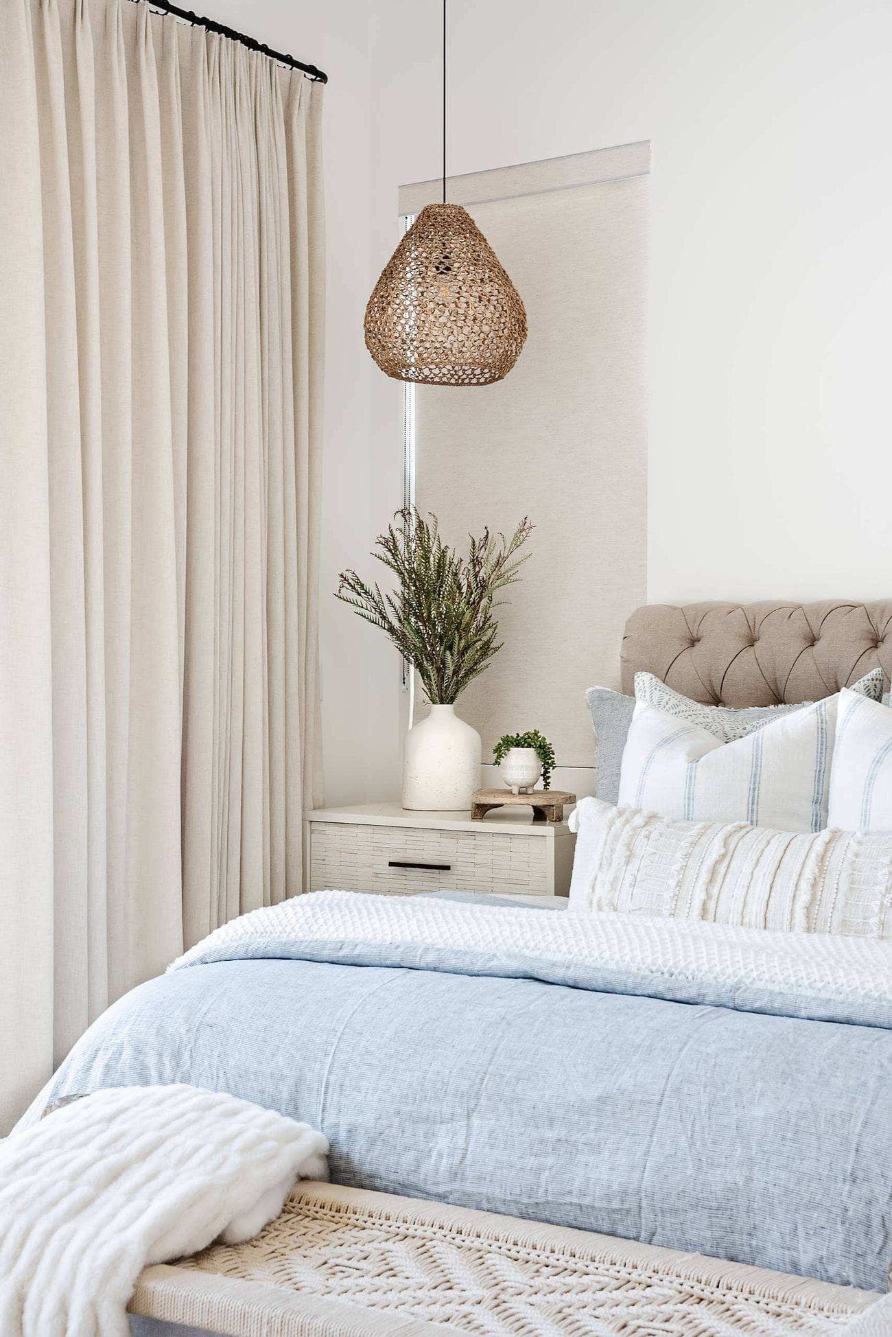 A linen-draped bedroom with a tufted headboard, a woven pendant and a washed-blue coverlet.