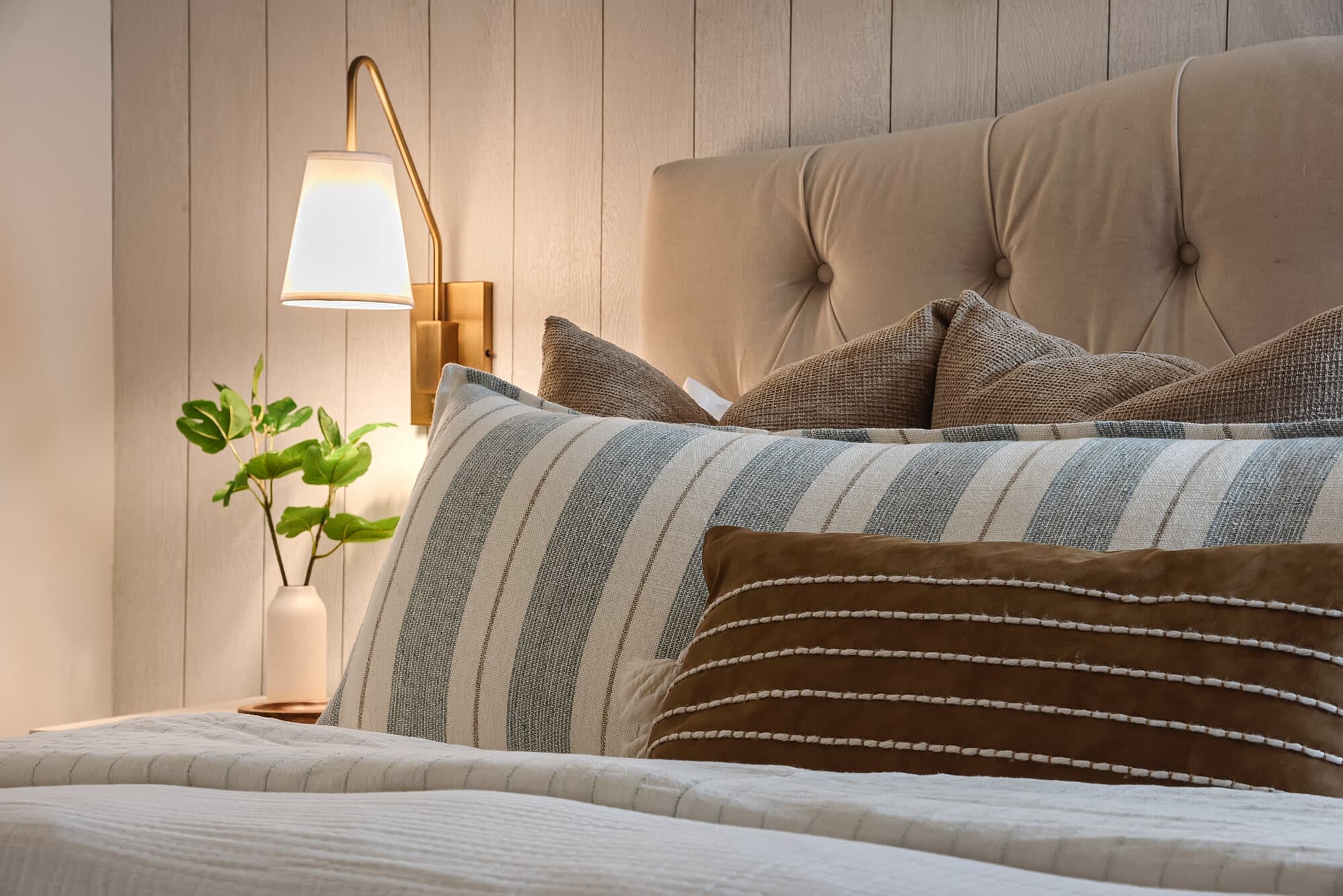 A made bed in linen and striped ticking with a dark velvet bolster, lit by a brass swing-arm sconce.